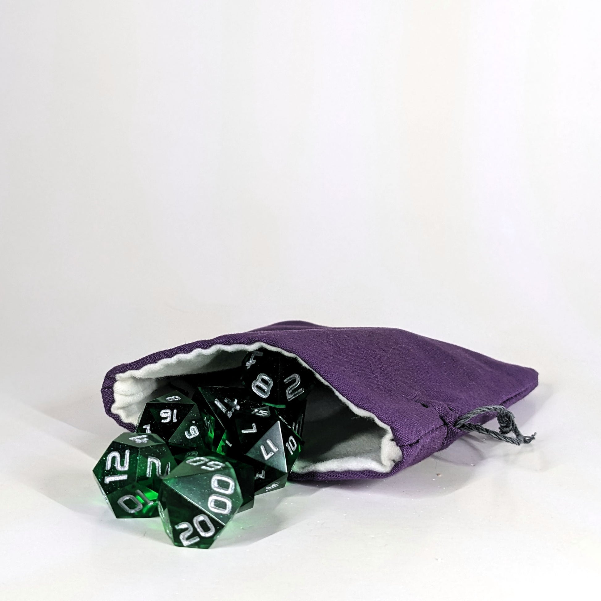 A small, handmade dice bag that is purple lays open with a set of green dnd dice with silver numbers spilling out of it on a white background.