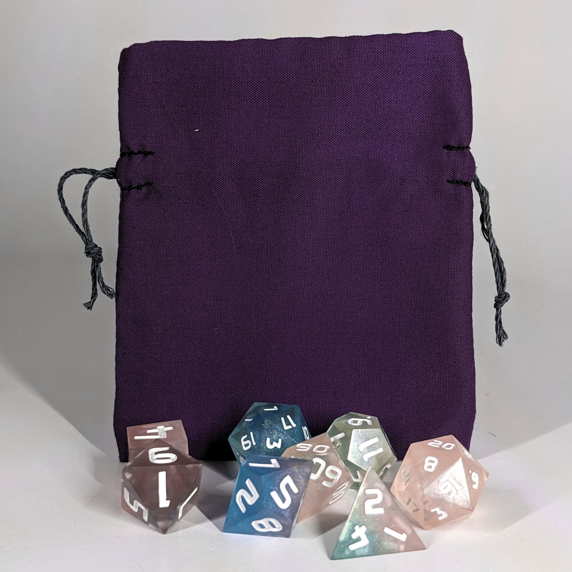 A small, handmade dice bag that is purple stands upright on a white background with a set of colorful dnd dice with white numbers sitting in front of it.