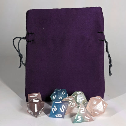 A small, handmade dice bag that is purple stands upright on a white background with a set of colorful dnd dice with white numbers sitting in front of it.