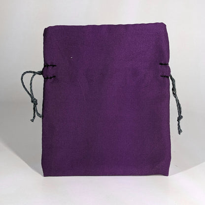 A small, handmade dice bag that is purple stands upright on a white background.