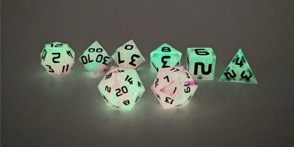 A straight on photo of a standard set of DnD dice that have pink/purple swirls showing through glow-in-the-dark green and black numbers focusing on 2 d20s in the center. The d20 on the left has the 20 face centered and the d20 on the right has the 1 face centered. The other 6 dice are lined up in the background.