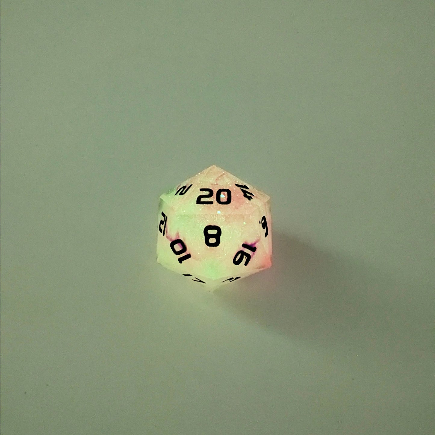 A high angle photo of a standard d20 glowing slightly green with pink/purple swirls and black numbers with the 20 face up on a white background.