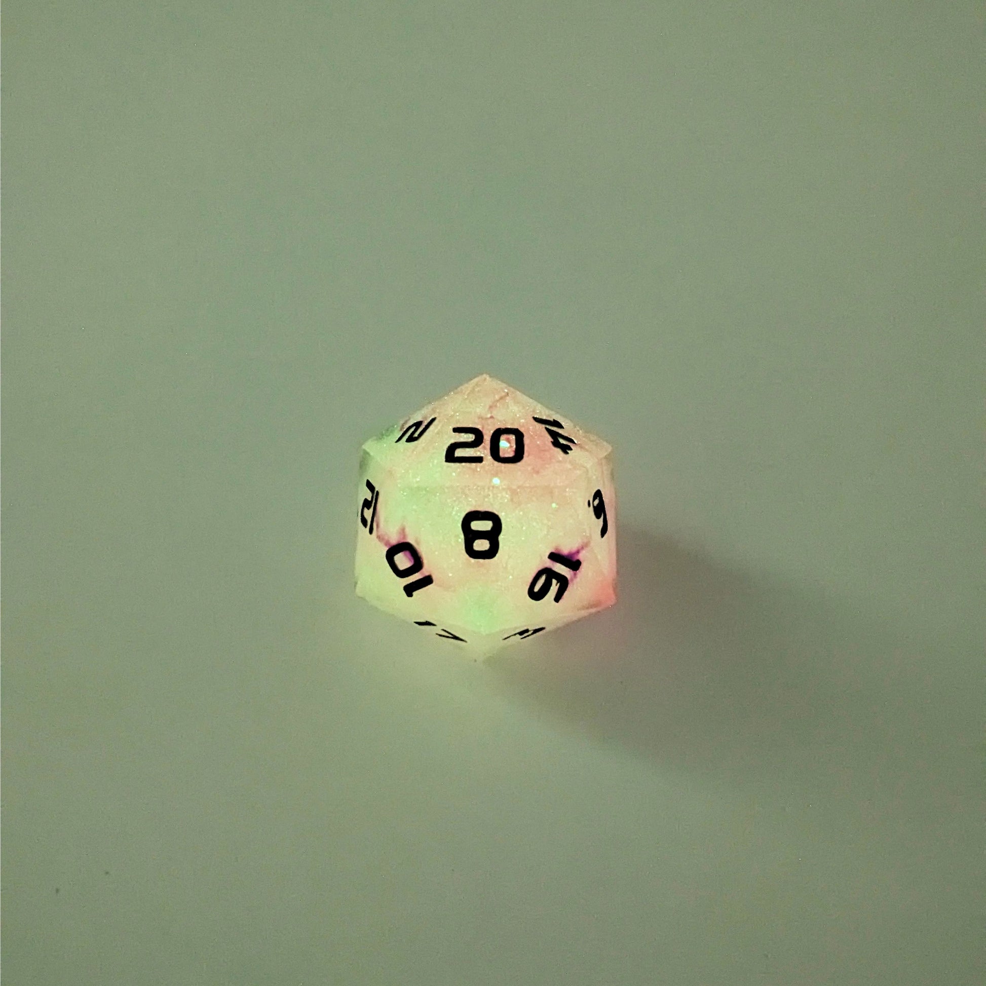 A high angle photo of a standard d20 glowing slightly green with pink/purple swirls and black numbers with the 20 face up on a white background.