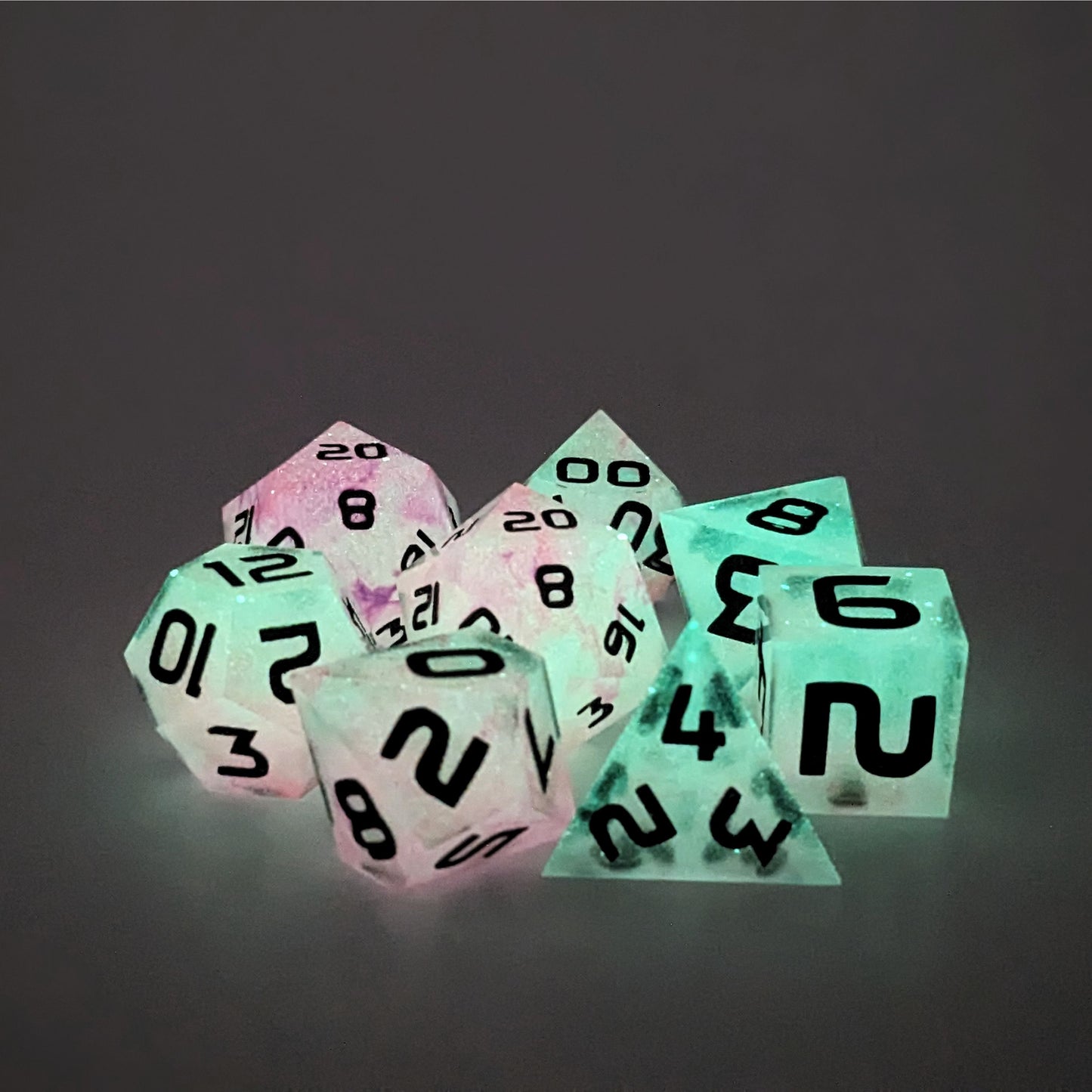A complete set of 8 polyhedral dice that are glowing green with pink/purple swirls and black numbers, showing the glow-in-the-dark feature of this dice set. The DnD dice set is arranged in a circle and features 2 D20s, one of which is in the center surrounded by the other 7 dice.