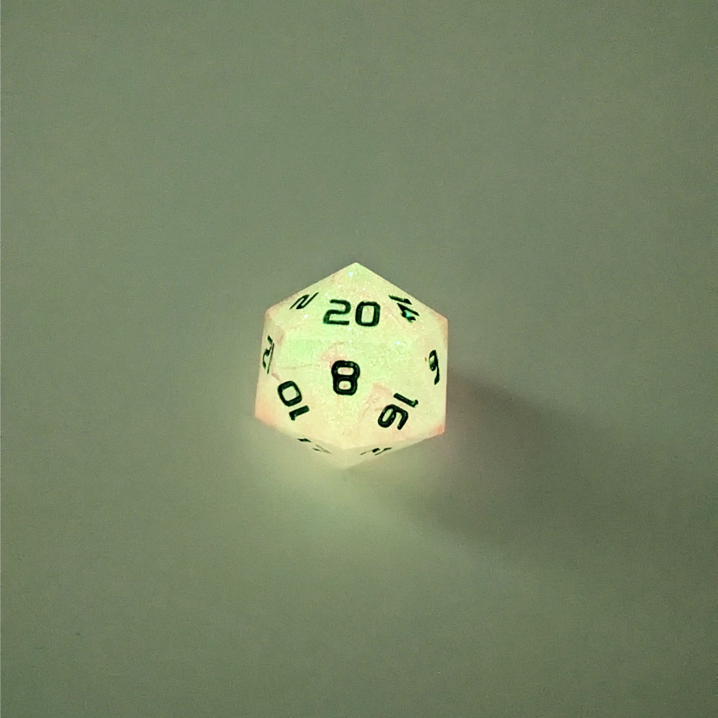 A high angle photo of a standard d20 glowing slightly green with pink/purple swirls and green numbers with the 20 face up on a white background.
