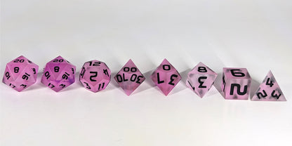 A straight on photo of a complete set of polyhedral dice that have pink/purple swirls and black numbers arranged in a straight line. They are lined up with the 2 d20s on the left going down to the d4 on the right side. The highest value number of each die is facing up.