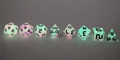 A straight on photo of a complete set of polyhedral dice that are glowing green with pink/purple swirls and have black numbers arranged in a straight line. They are lined up with the 2 d20s on the left going down to the d4 on the right side. The highest value number of each die is facing up.