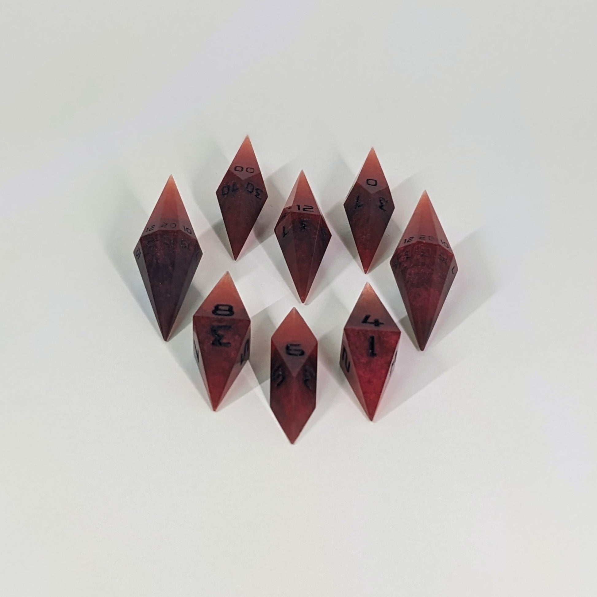 A complete set of resin dice featuring 2 d20s for advantage rolls. These mixed shades of red with black numbering slender handmade diamond dice are clustered in a tight circle on a white background, highlighting the quality of the artisan dice craftsmanship and the unique pointed shape.
