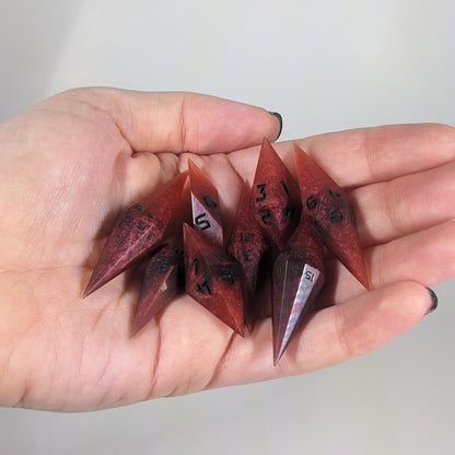 Close-up of mixed shades of red with black numbering artisan dice resting in a palm on a seamless white backdrop. The handmade dice feature a distinct sharp diamond shape, with 2 d20s visible among the high-quality resin dice.