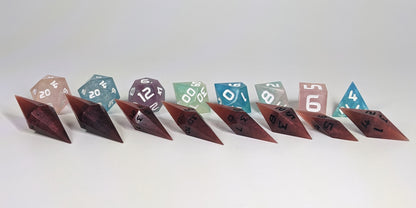 A size comparison of mixed shades of red with black numbering long pointed diamond handmade dice with standard shapes placed behind for scale. The photo emphasizes the length of the diamond dice being much greater than a standard set as well as the diamond set being much more slender. This artisan dice set features 2 d20s and rests on a seamless white background to highlight the elongated profile of the resin dice.