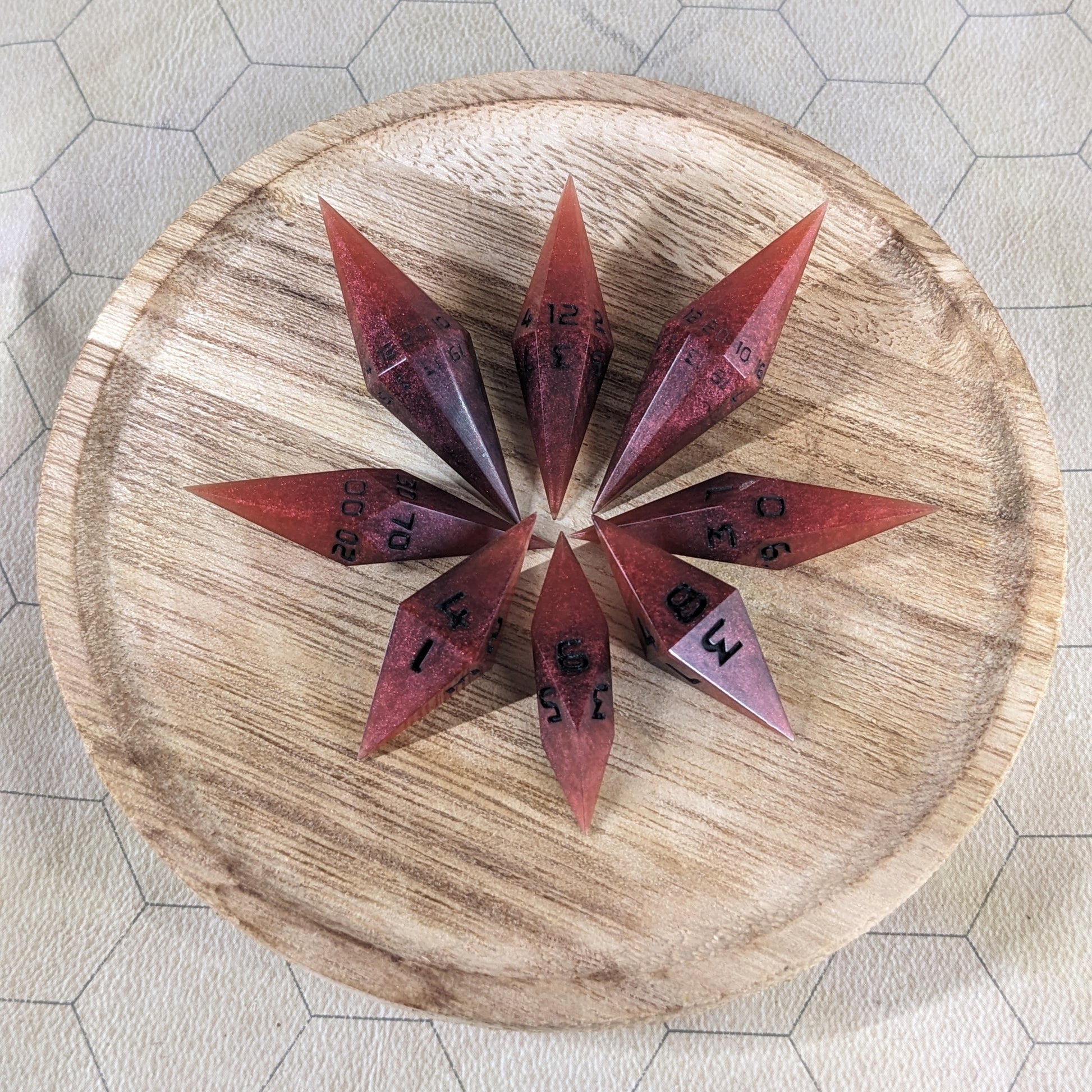 A set of mixed shades of red with black numbering handmade dice arranged in a wooden circle organizer. The resin dice feature a unique pointed diamond shape with the tips facing outward, highlighting the sharp edges of the two d20 and other artisan dice in the collection. 