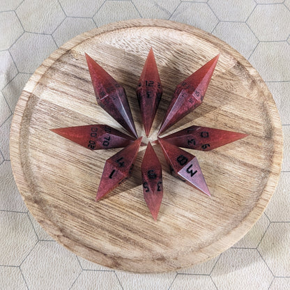 A set of mixed shades of red with black numbering handmade dice arranged in a wooden circle organizer. The resin dice feature a unique pointed diamond shape with the tips facing outward, highlighting the sharp edges of the two d20 and other artisan dice in the collection. 