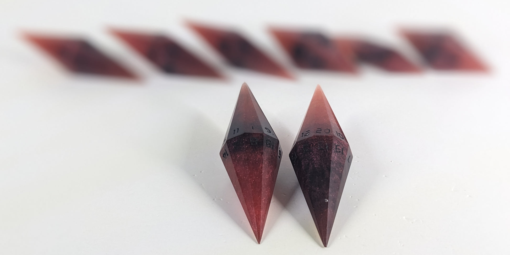 A stylized macro shot focusing heavily on 2 d20s from a mixed shades of red with black numbering handmade dice set in the foreground. The sharp diamond edges of these front resin dice are crisp, while the rest of the matching artisan dice collection forms a softly blurred background, creating depth.