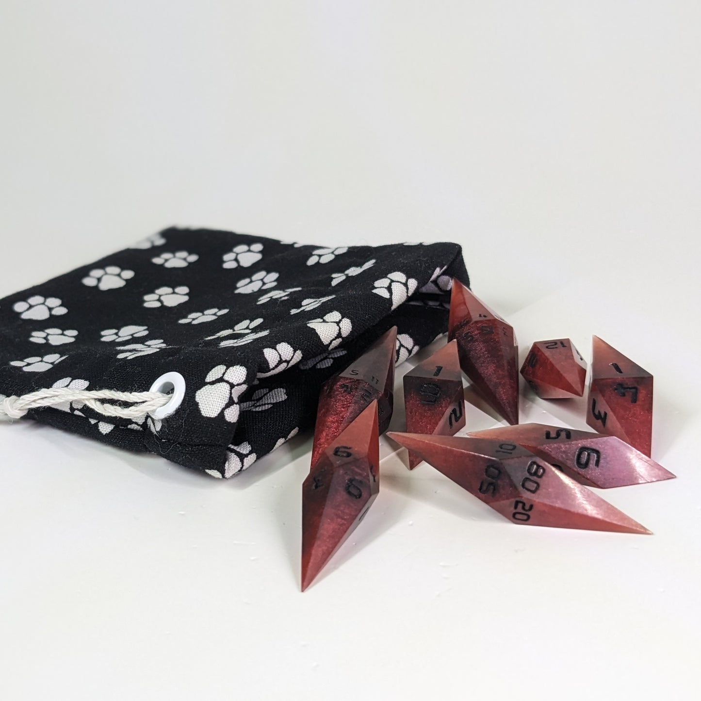 Mixed shades of red with black numbering diamond handmade dice spilling out of a (color description) drawstring bag on a white background. The set features 2 d20s and matching resin dice, showcasing the unique shape of these artisan dice.