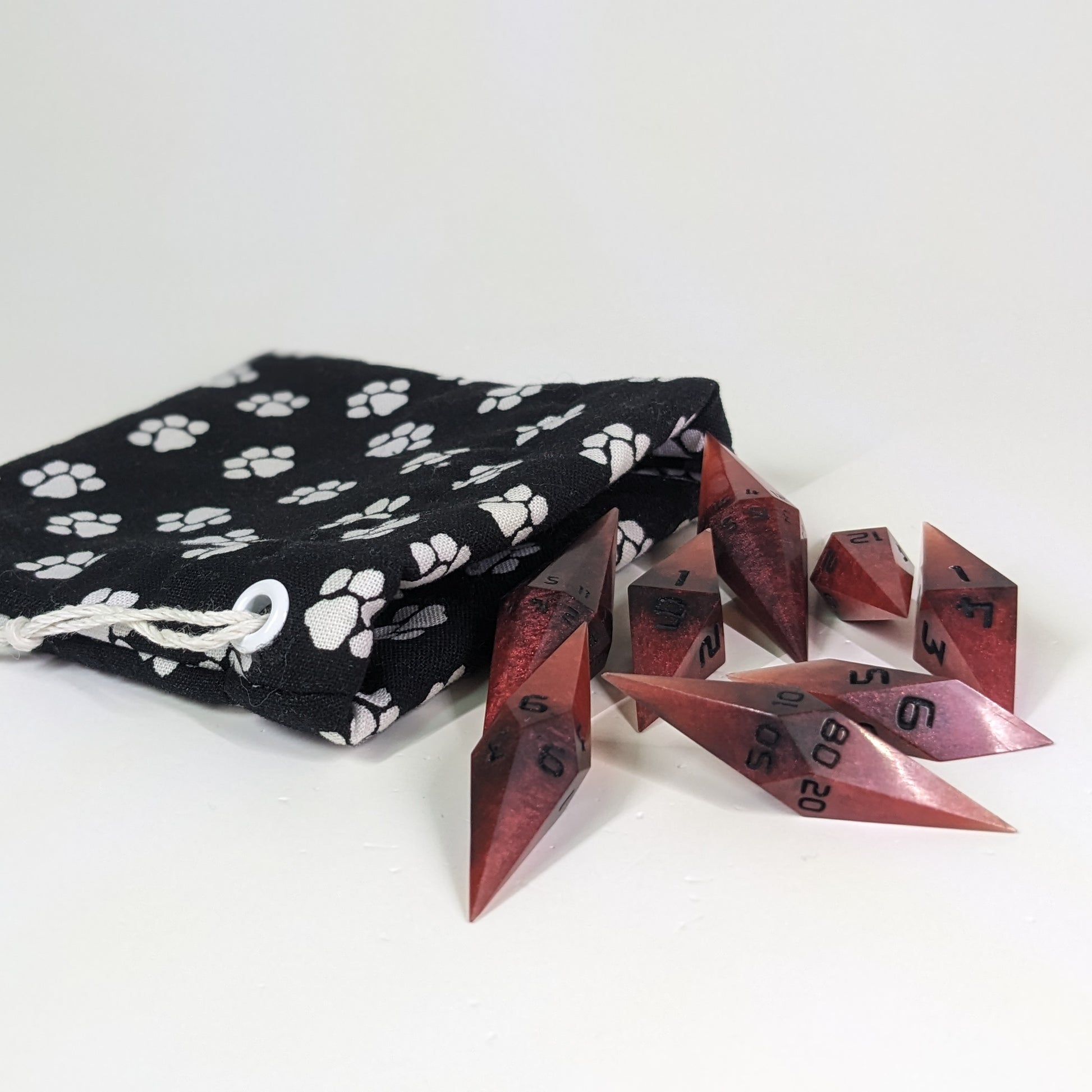 Mixed shades of red with black numbering diamond handmade dice spilling out of a (color description) drawstring bag on a white background. The set features 2 d20s and matching resin dice, showcasing the unique shape of these artisan dice.