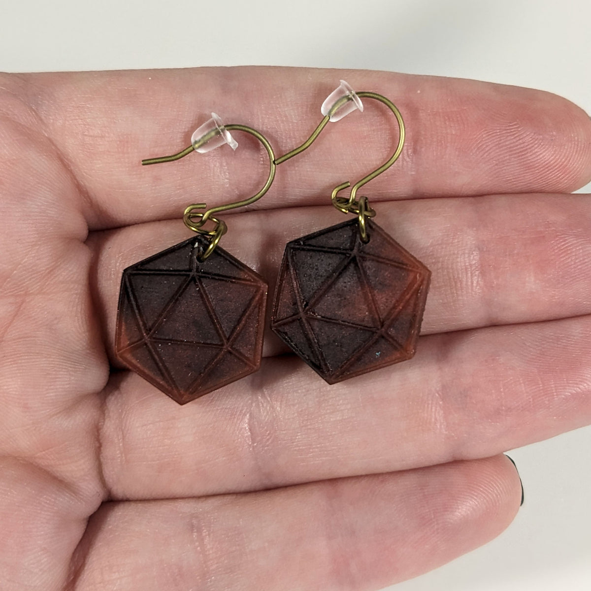 A pair of maroon and red dangly, flat d20 earrings with gold color titanium and niobium hardware with bell shaped rubber backs held on on a hand on a white background.