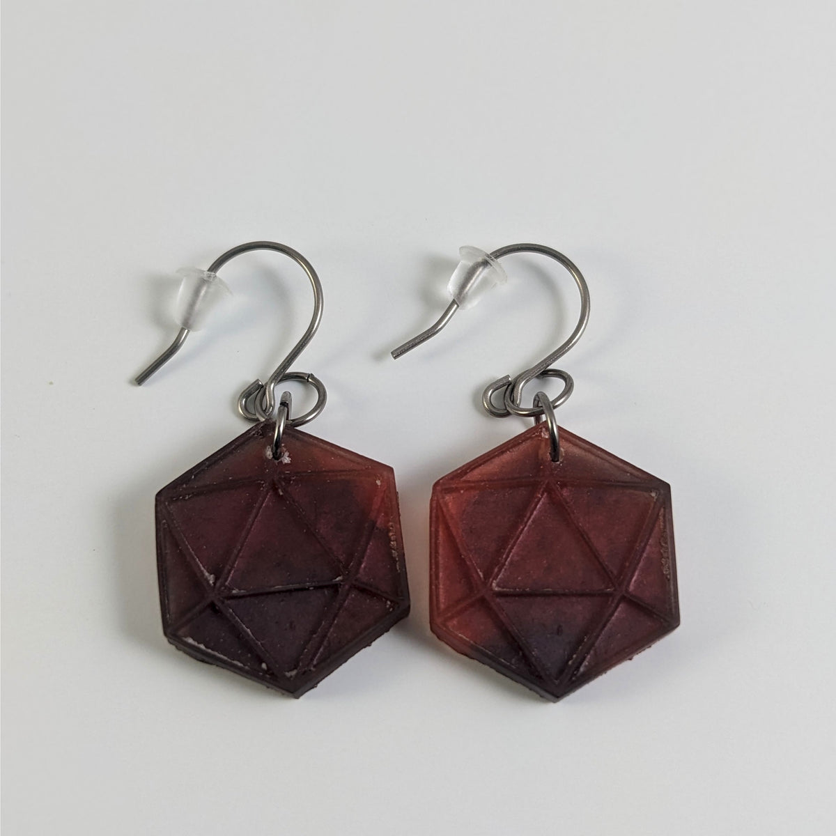 A pair of maroon and red dangly, flat d20 earrings with silver color titanium and niobium hardware and bell shaped rubber backs on a white background.