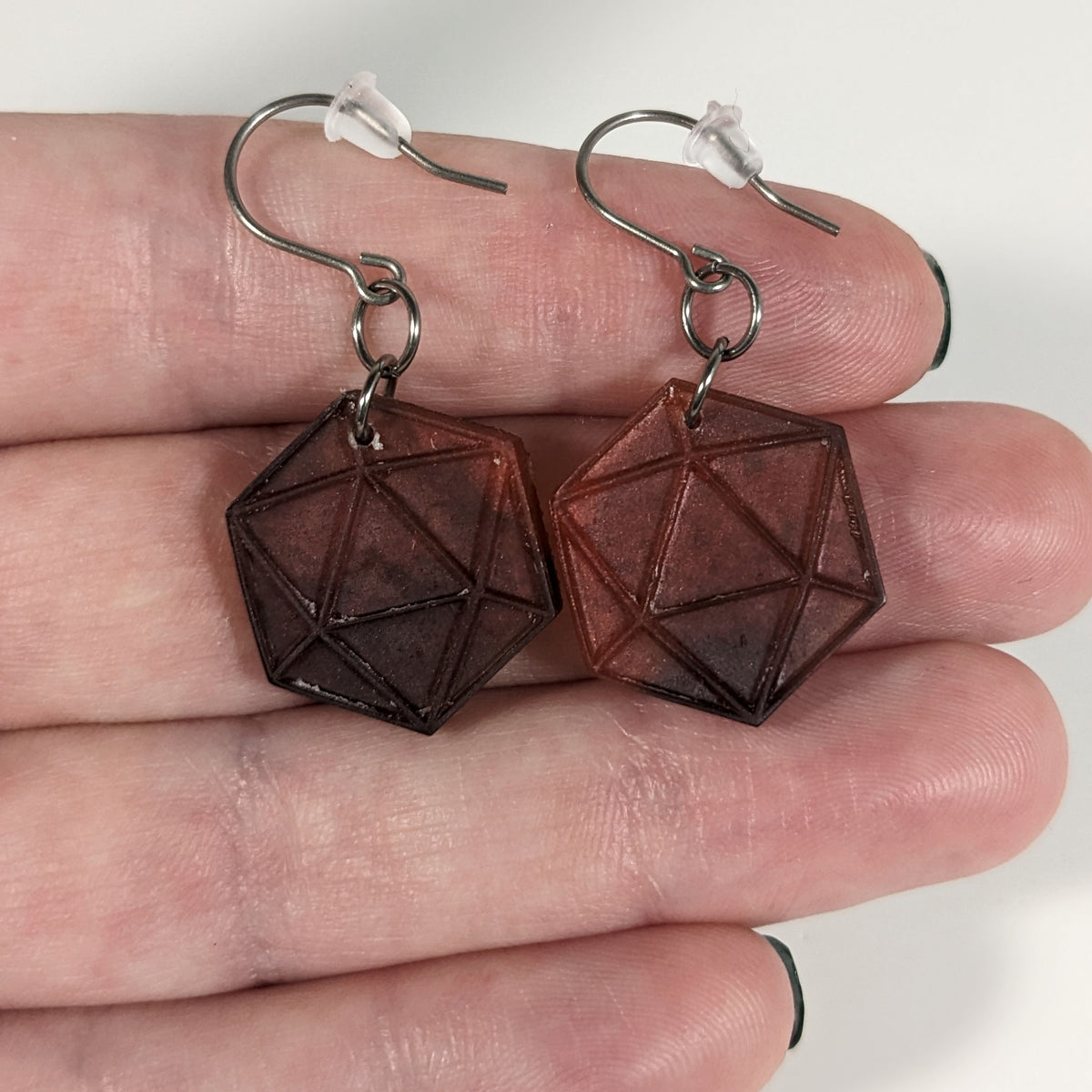 A pair of maroon and red dangly, flat d20 earrings with silver color titanium and niobium hardware with bell shaped rubber backs held on on a hand on a white background.