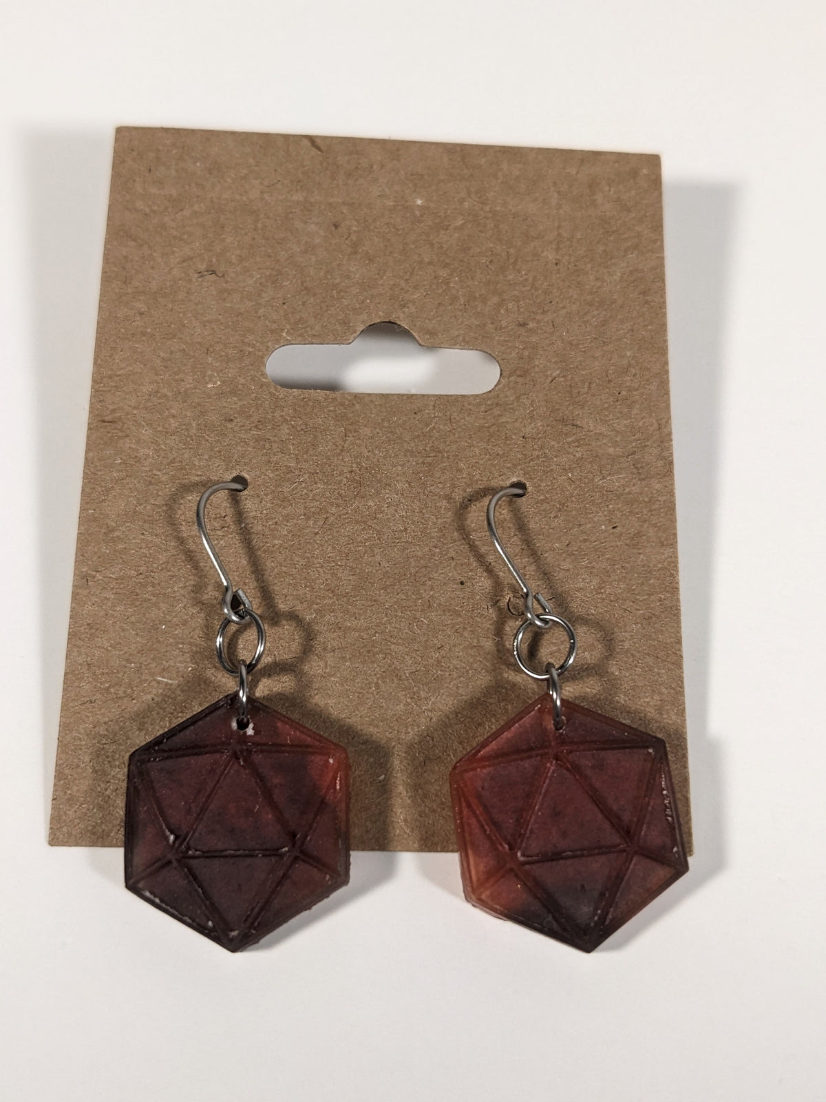 A pair of maroon and red dangly, flat d20 earrings with silver color titanium and niobium hardware on a kraft earring card on a white background.