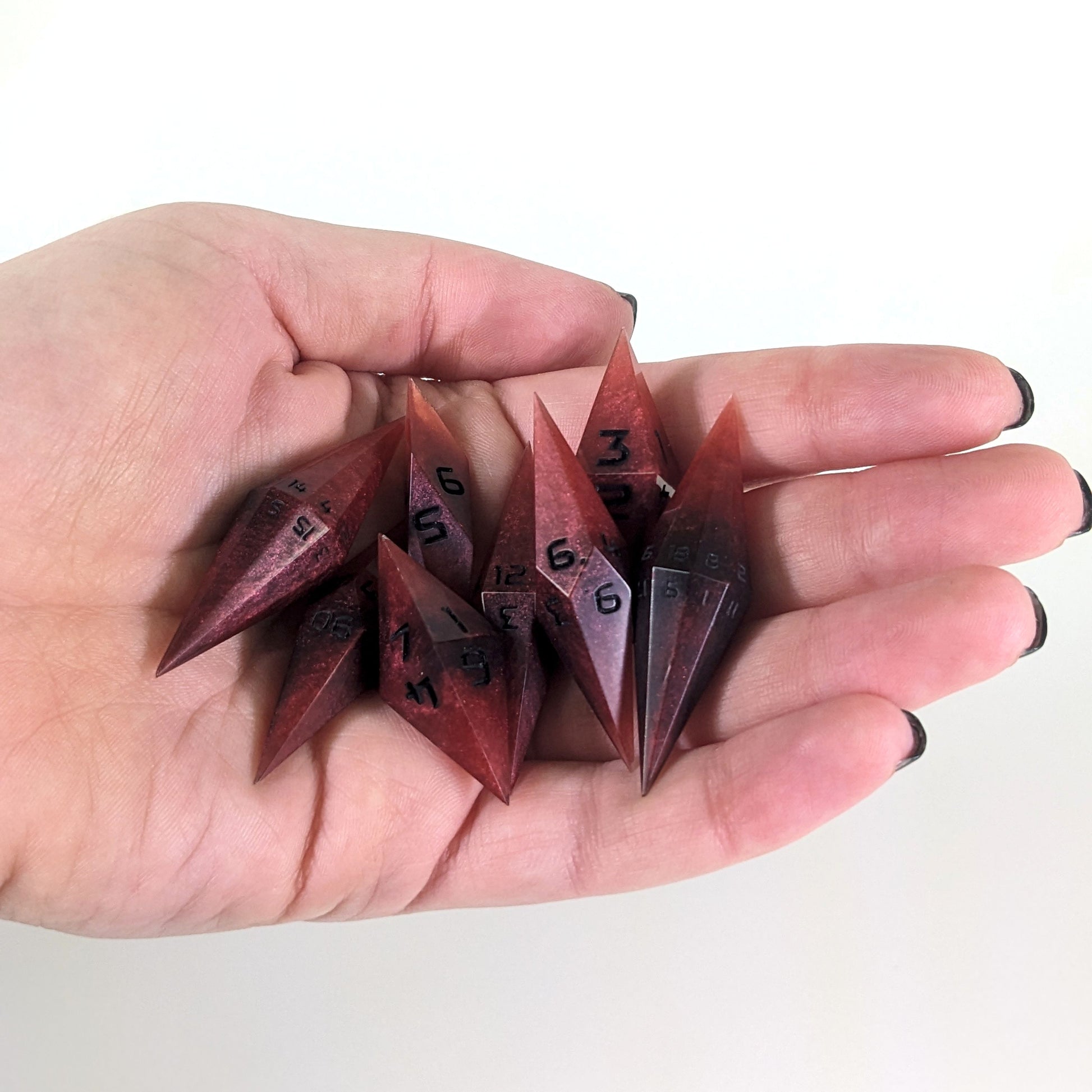 An 8 dice set of diamond style dice that are mixed shades of red with black numbers are held in a hand. The image shows that the set fits easily in a hand, making them overall smaller, though sharper, than a standard set of dnd dice.