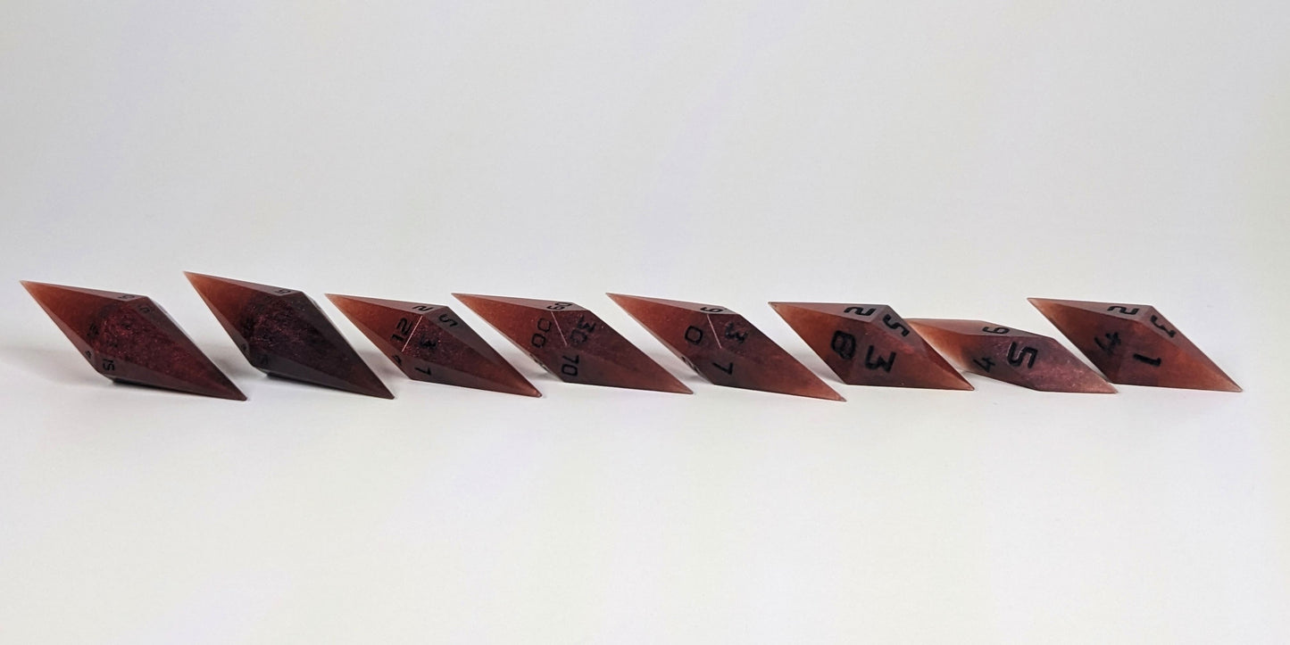 A mid angle photo of a complete set of handmade diamond DnD dice that are mixed shades of red with black numbers arranged in a straight line with bottom points angled slightly forwards and towards the right. They are lined up with the 2 d20s on the left going down to the d4 on the right side. The highest value number of each die is facing forward.
