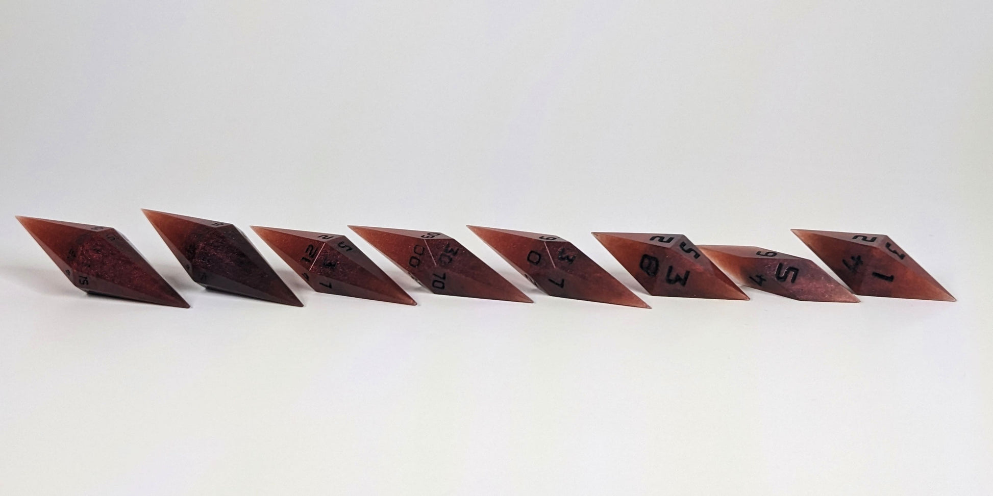 A mid angle photo of a complete set of handmade diamond DnD dice that are mixed shades of red with black numbers arranged in a straight line with bottom points angled slightly forwards and towards the right. They are lined up with the 2 d20s on the left going down to the d4 on the right side. The highest value number of each die is facing forward.
