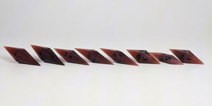 A mid angle photo of a complete set of handmade diamond DnD dice that are mixed shades of red with black numbers arranged in a straight line with bottom points angled slightly forwards and towards the right. They are lined up with the 2 d20s on the left going down to the d4 on the right side. The highest value number of each die is facing forward.