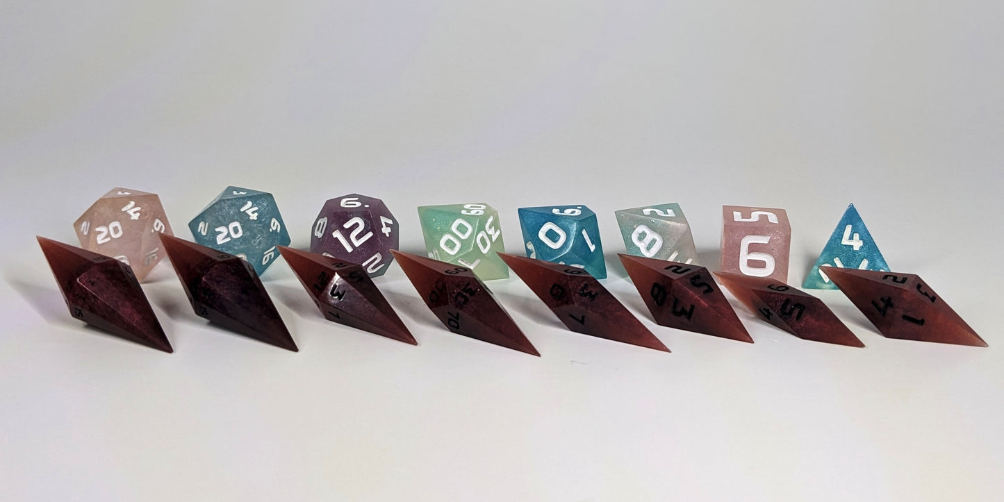 A straight on photo of a complete set of dagger dice that are mixed shades of red with black numbers arranged in a straight line with points angled forward and to the right. They are lined up with the 2 d20s on the left going down to the d4 on the right side. Behind the dagger set is a standard set of polyhedral dice to allow for size comparison. The diamond dice are much smaller overall. The highest value number of each die is facing forward.