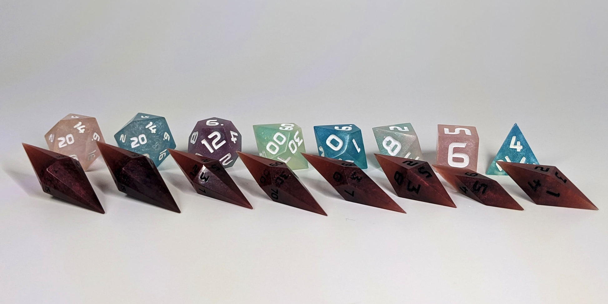 A straight on photo of a complete set of dagger dice that are mixed shades of red with black numbers arranged in a straight line with points angled forward and to the right. They are lined up with the 2 d20s on the left going down to the d4 on the right side. Behind the dagger set is a standard set of polyhedral dice to allow for size comparison. The diamond dice are much smaller overall. The highest value number of each die is facing forward.