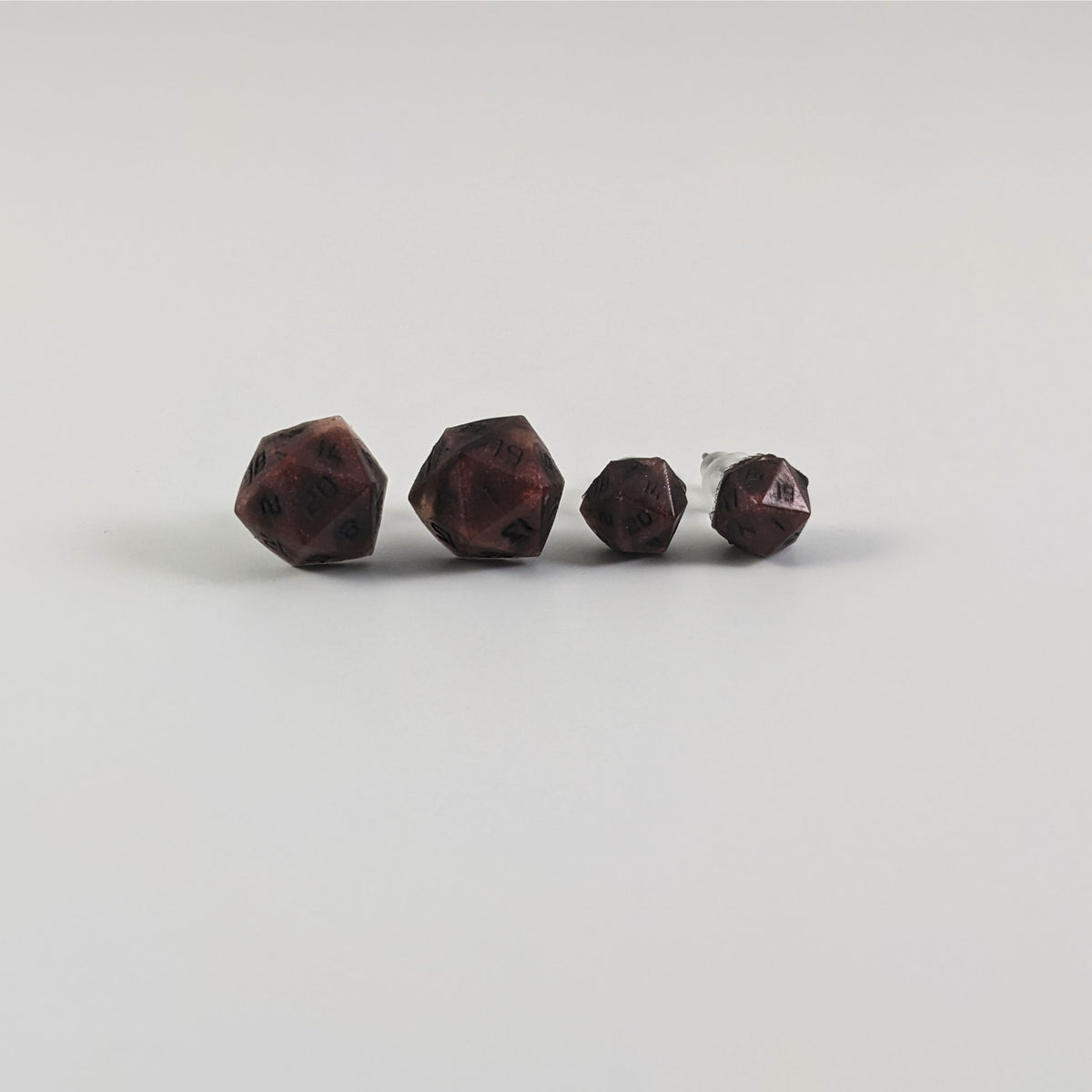 Two pairs of handmade d20 earrings with titanium hardware that are swirls of red with black numbers on a white background sit in a line with the larger pair on the left and the smaller pair on the right. The earrings on the left of each pair centers the 20 face and the earrings on the right of each pair centers the 1 face.
