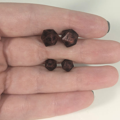 Two pairs of handmade d20 earrings with titanium hardware that are swirls of red with black numbers held between the fingers of a flat hand. The larger pair is between the pointer and middle finger, the smaller between the middle and ring finger. The earrings on the left of each pair centers the 20 face and the earrings on the right of each pair centers the 1 face.