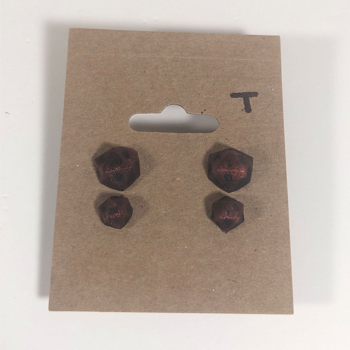 Two pairs of handmade d20 earrings with titanium hardware that are swirls of red with black numbers on a kraft brown earring card with a T in the top right corner. The earrings on the left of each pair centers the 20 face and the earrings on the right of each pair centers the 1 face.