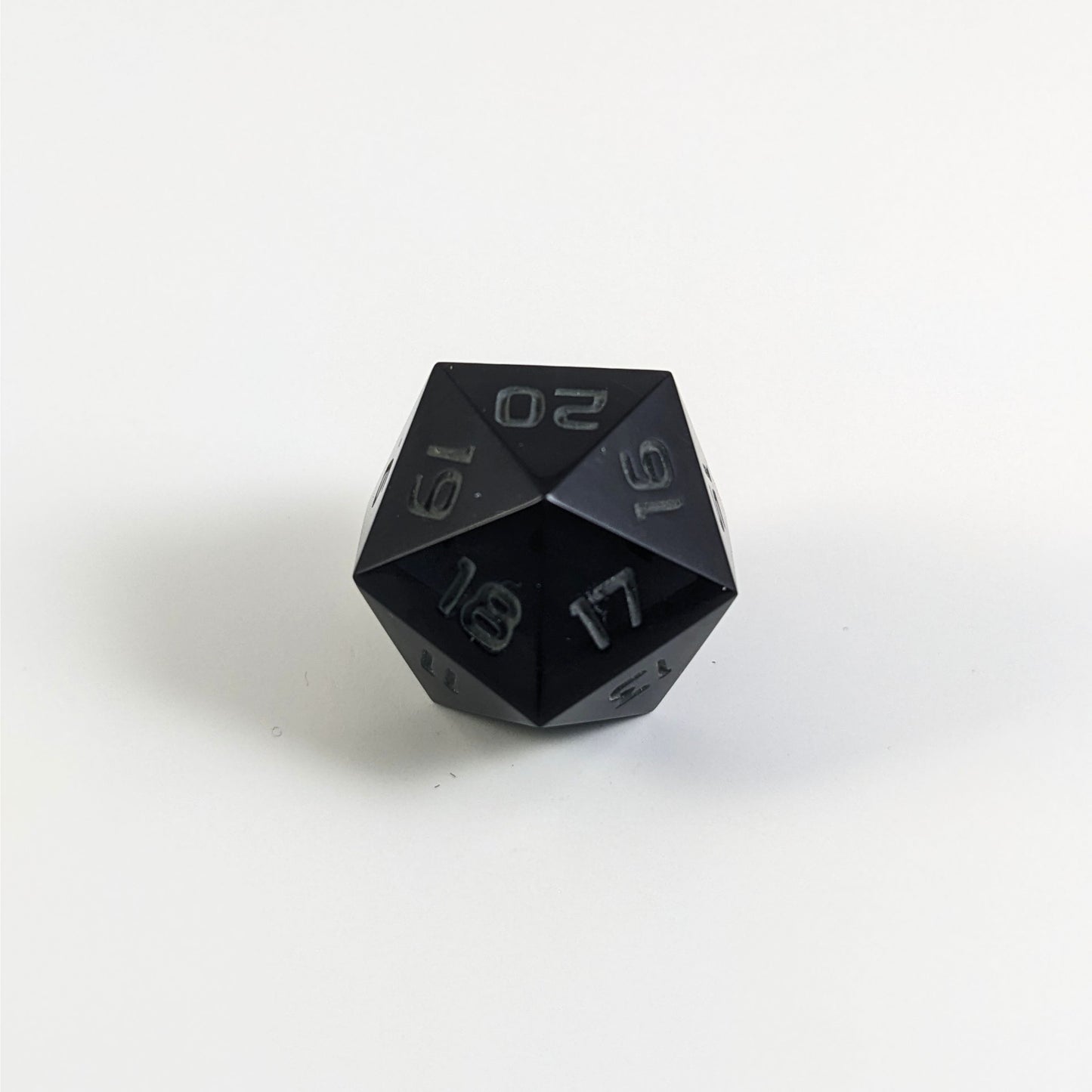 A low angle of a black spindown d20 with gray numbers and a right spin sits on a white background with the 20 through 16 corner facing the camera showing the spiral of the spin down.