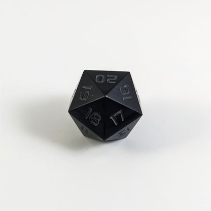A low angle of a black spindown d20 with gray numbers and a right spin sits on a white background with the 20 through 16 corner facing the camera showing the spiral of the spin down.
