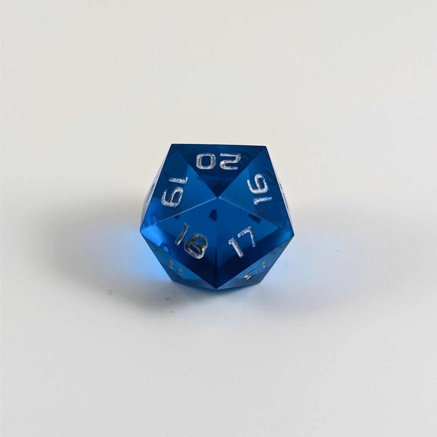 A low angle of a blue spindown d20 with silver numbers and a right spin sits on a white background with the 20 through 16 corner facing the camera showing the spiral of the spin down.