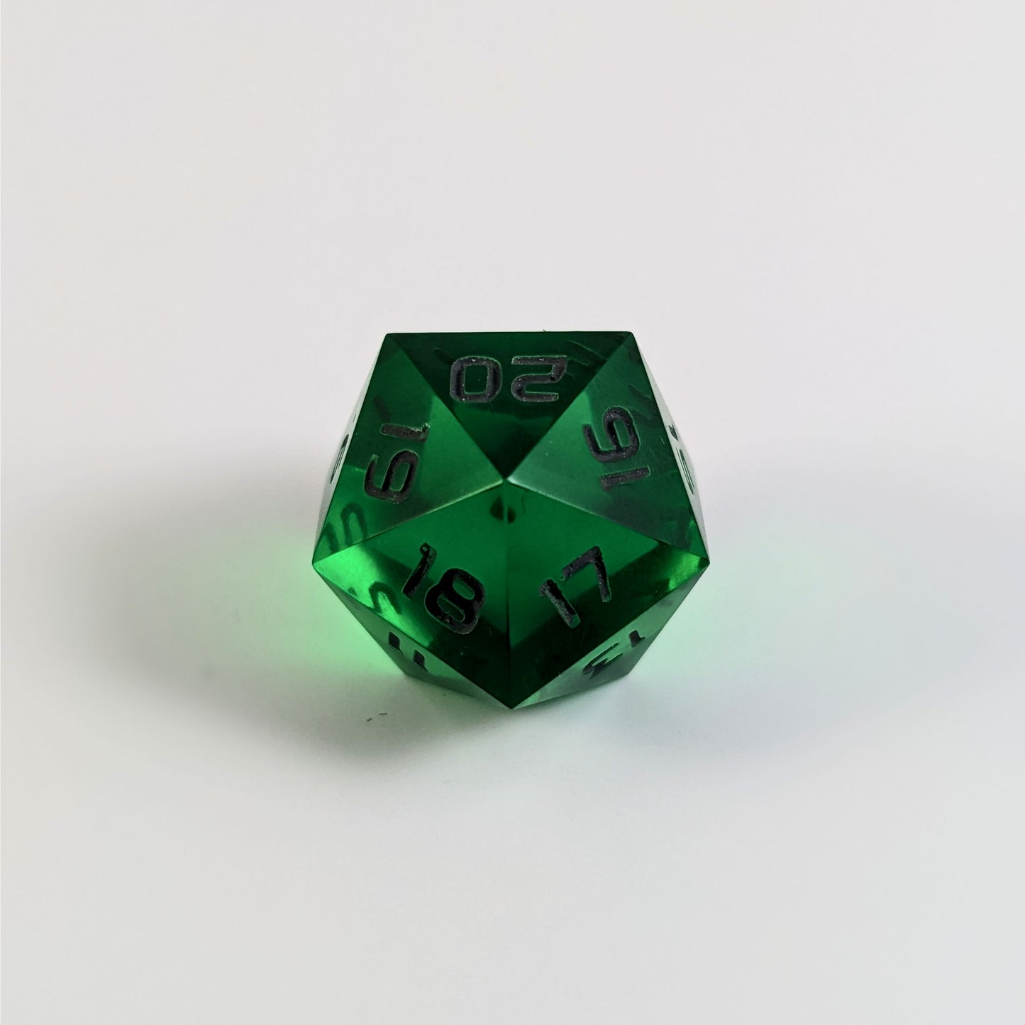 A low angle of a green spindown d20 with gray numbers and a right spin sits on a white background with the 20 through 16 corner facing the camera showing the spiral of the spin down.