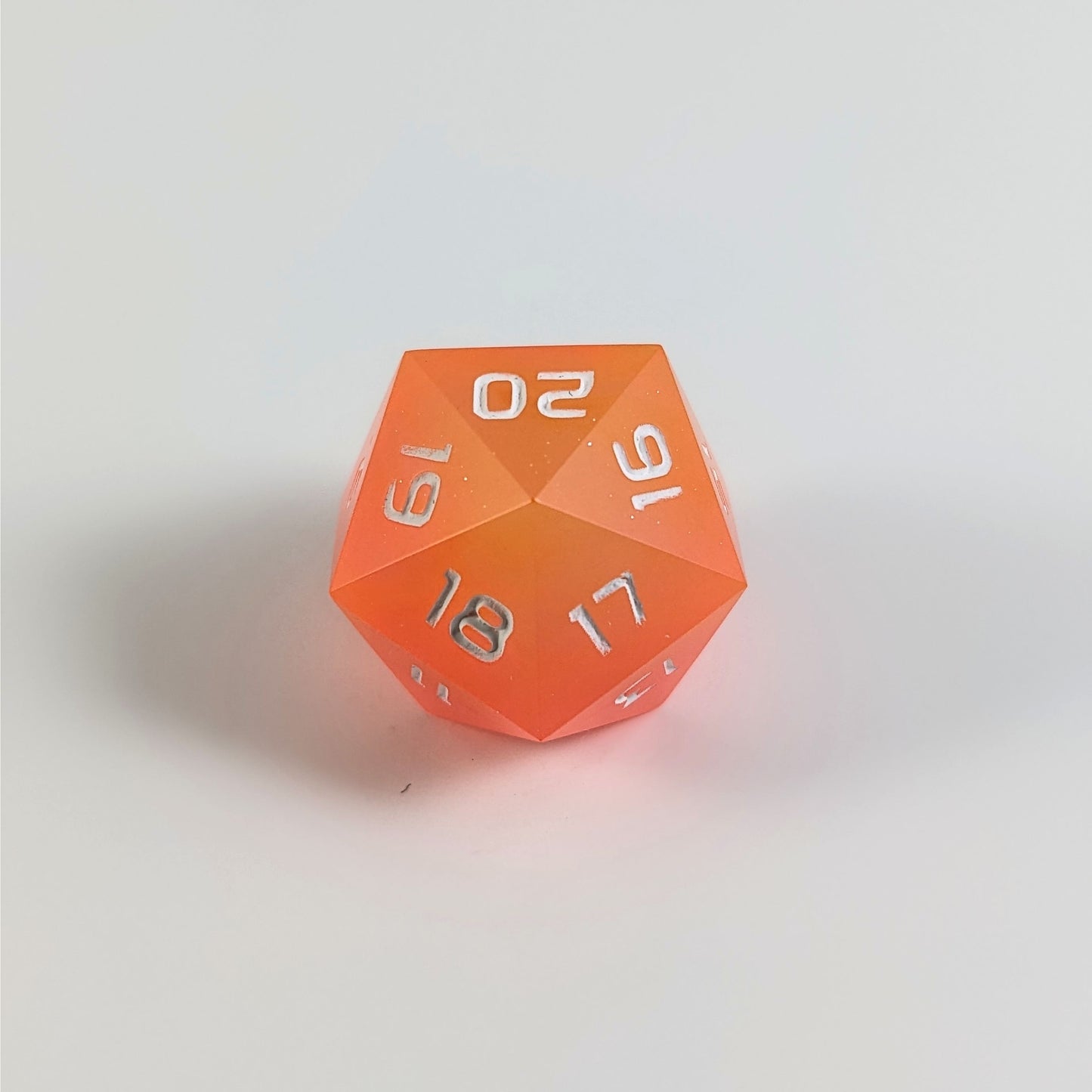 A low angle of an orange spindown d20 with white numbers and a right spin sits on a white background with the 20 through 16 corner facing the camera showing the spiral of the spin down.