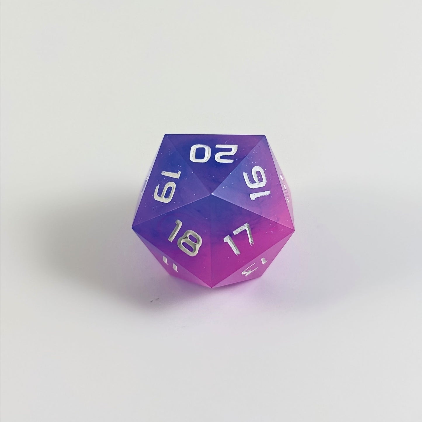 A low angle of a pink and blue spindown d20 with white numbers and a right spin sits on a white background with the 20 through 16 corner facing the camera showing the spiral of the spin down.