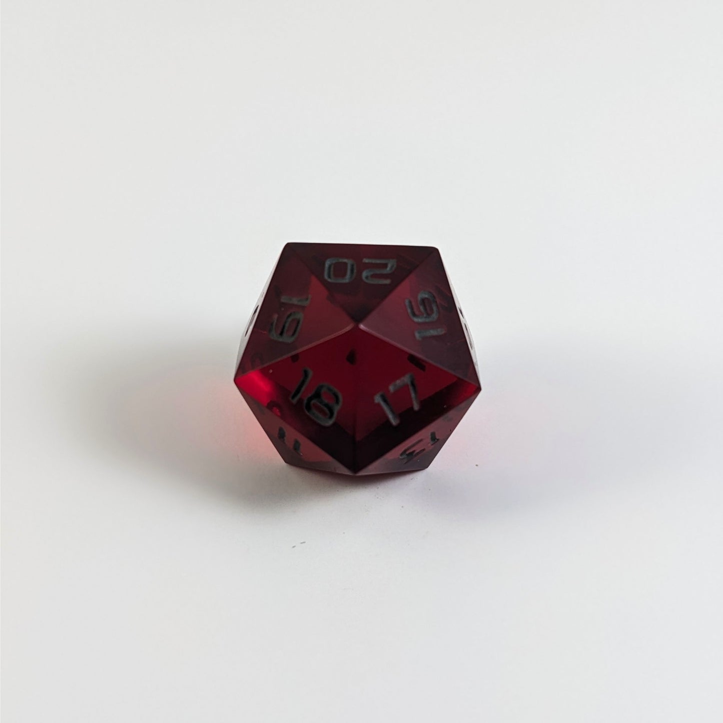 A low angle of a red spindown d20 with gray numbers and a right spin sits on a white background with the 20 through 16 corner facing the camera showing the spiral of the spin down.