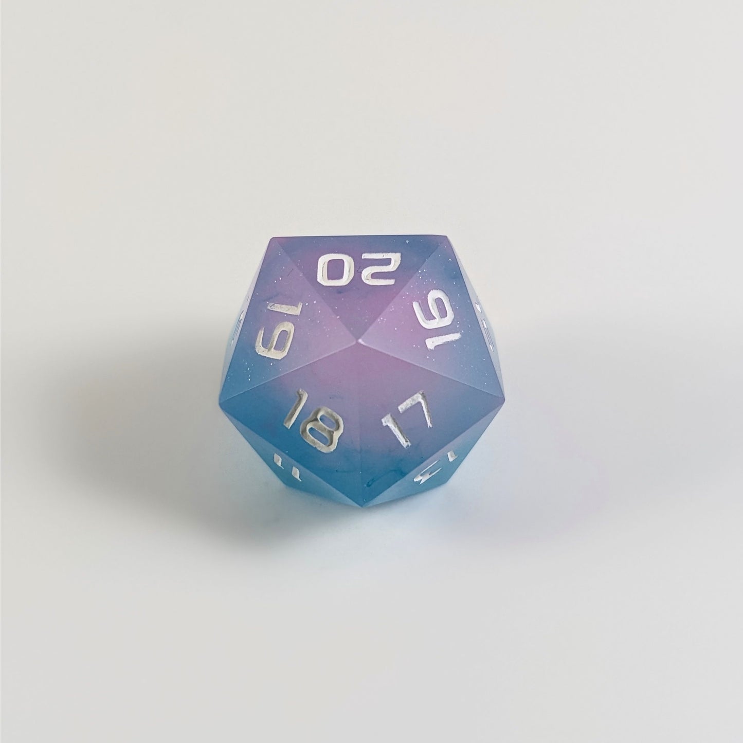 A low angle of an opaque pink and teal spindown d20 with white numbers and a right spin sits on a white background with the 20 through 16 corner facing the camera showing the spiral of the spin down.
