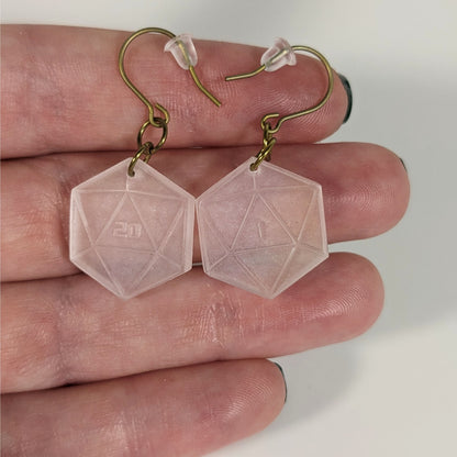 A pair of pale pink and white dangly, flat d20 earrings with gold color titanium and niobium hardware with bell shaped rubber backs held on on a hand on a white background. The earring on the left has a 20 on it and the earring on the right has a 1 on it, both in a sci-fi style font.
