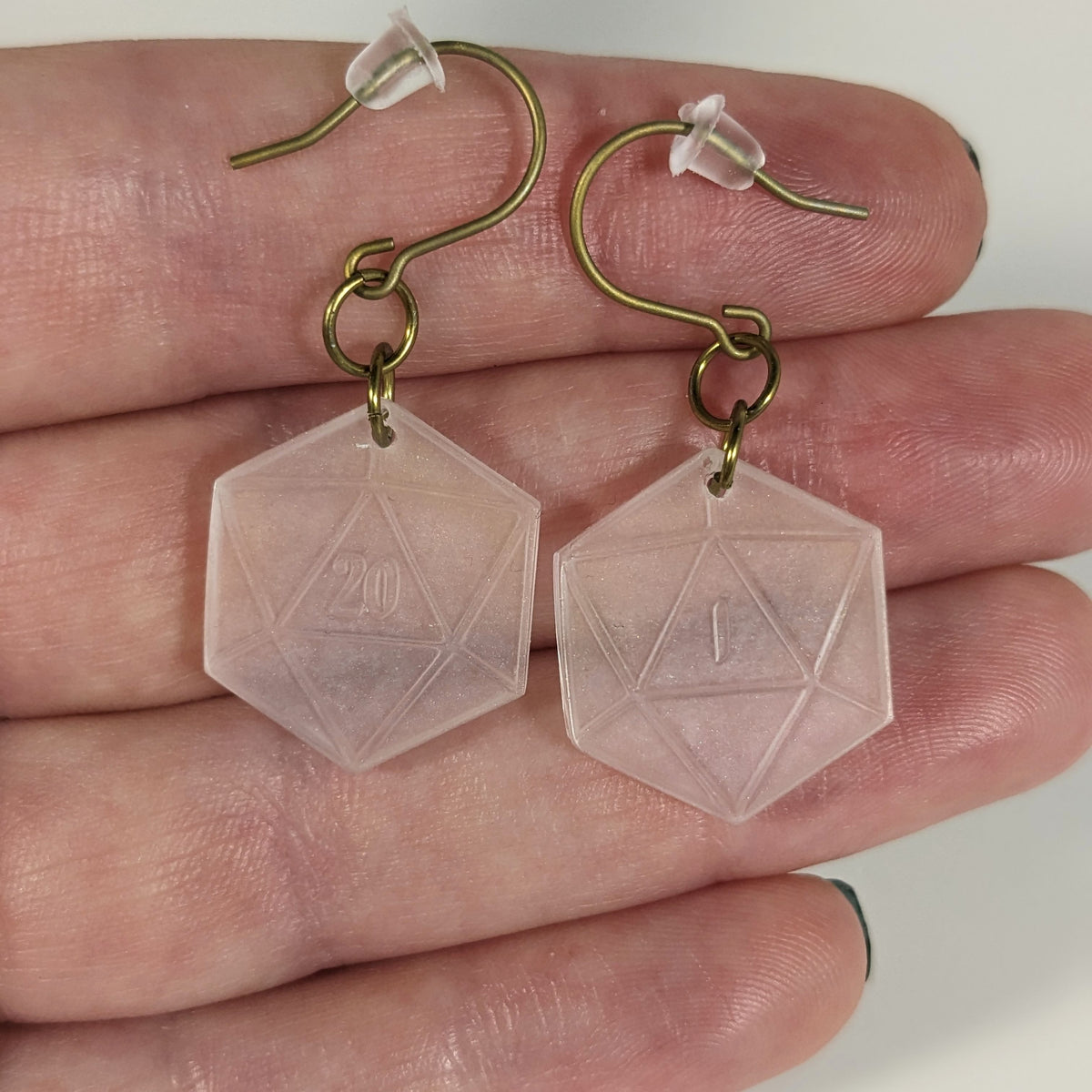 A pair of pale pink and white dangly, flat d20 earrings with gold color titanium and niobium hardware with bell shaped rubber backs held on on a hand on a white background. The earring on the left has a 20 on it and the earring on the right has a 1 on it, both in a fantasy style font.