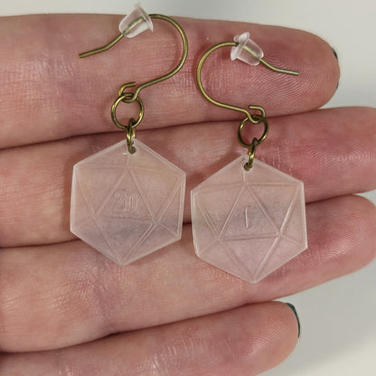 A pair of pale pink and white dangly, flat d20 earrings with gold color titanium and niobium hardware with bell shaped rubber backs held on on a hand on a white background. The earring on the left has a 20 on it and the earring on the right has a 1 on it, both in a fantasy style font.