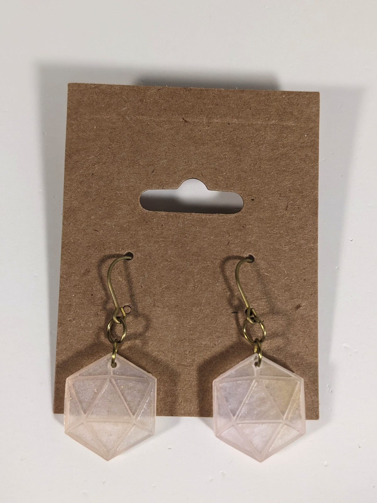 A pair of pale pink and white dangly, flat d20 earrings with gold color titanium and niobium hardware on a kraft earring card on a white background.