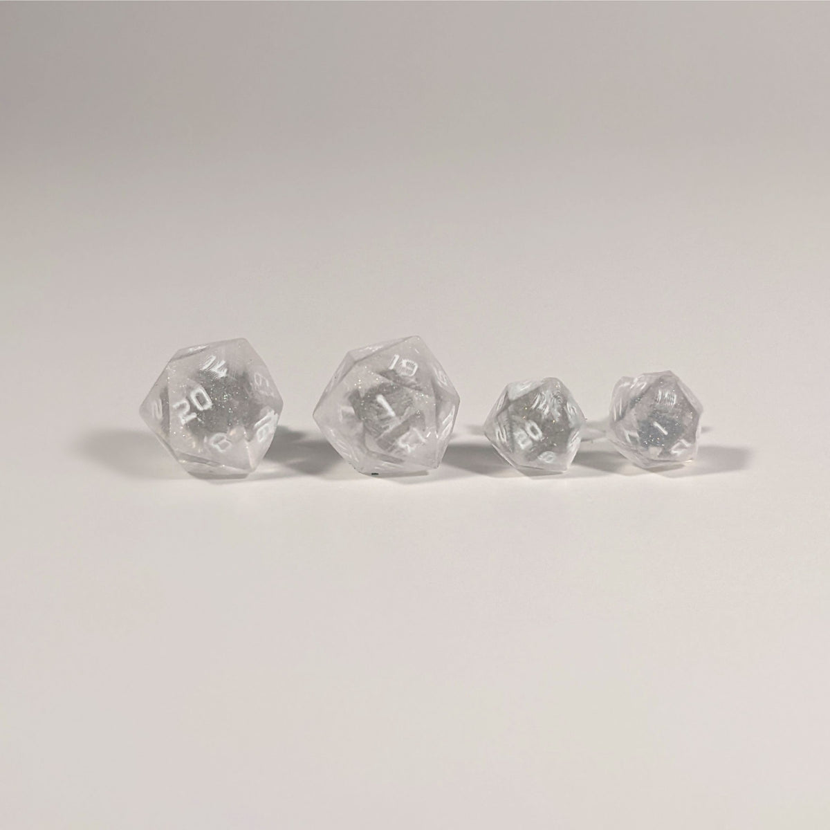 Two pairs of handmade d20 earrings with titanium hardware that are faded pink and translucent white with white numbers on a white background sit in a line with the larger pair on the left and the smaller pair on the right. The earrings on the left of each pair centers the 20 face and the earrings on the right of each pair centers the 1 face.