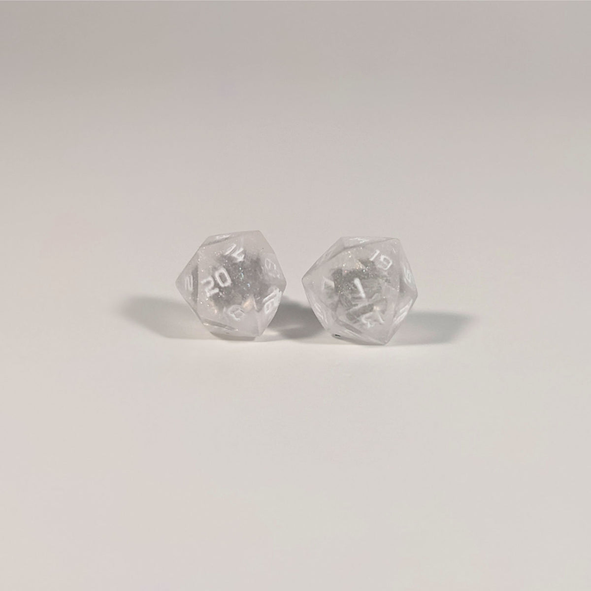 A single pair of the larger size handmade d20 earrings with titanium hardware that are glittery clear with pink flakies with white numbers on a white background. The earring on the left centers the 20 face and the earring on the right centers the 1 face.