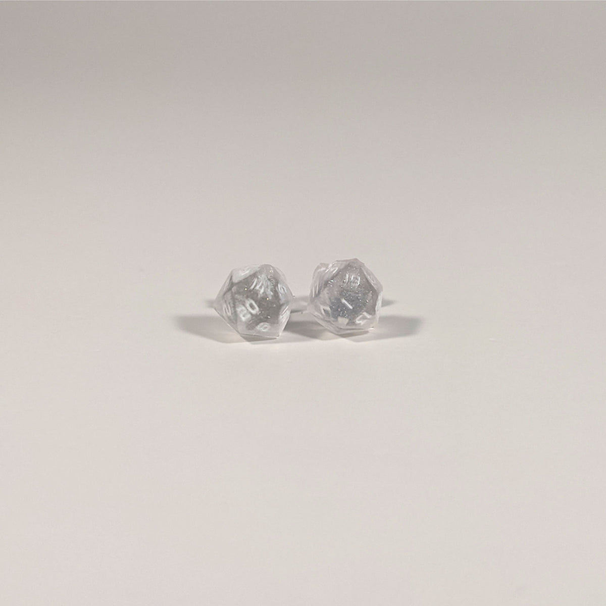A single pair of the smaller size handmade d20 earrings with titanium hardware that are glittery clear with pink flakies with white numbers on a white background. The earring on the left centers the 20 face and the earring on the right centers the 1 face.