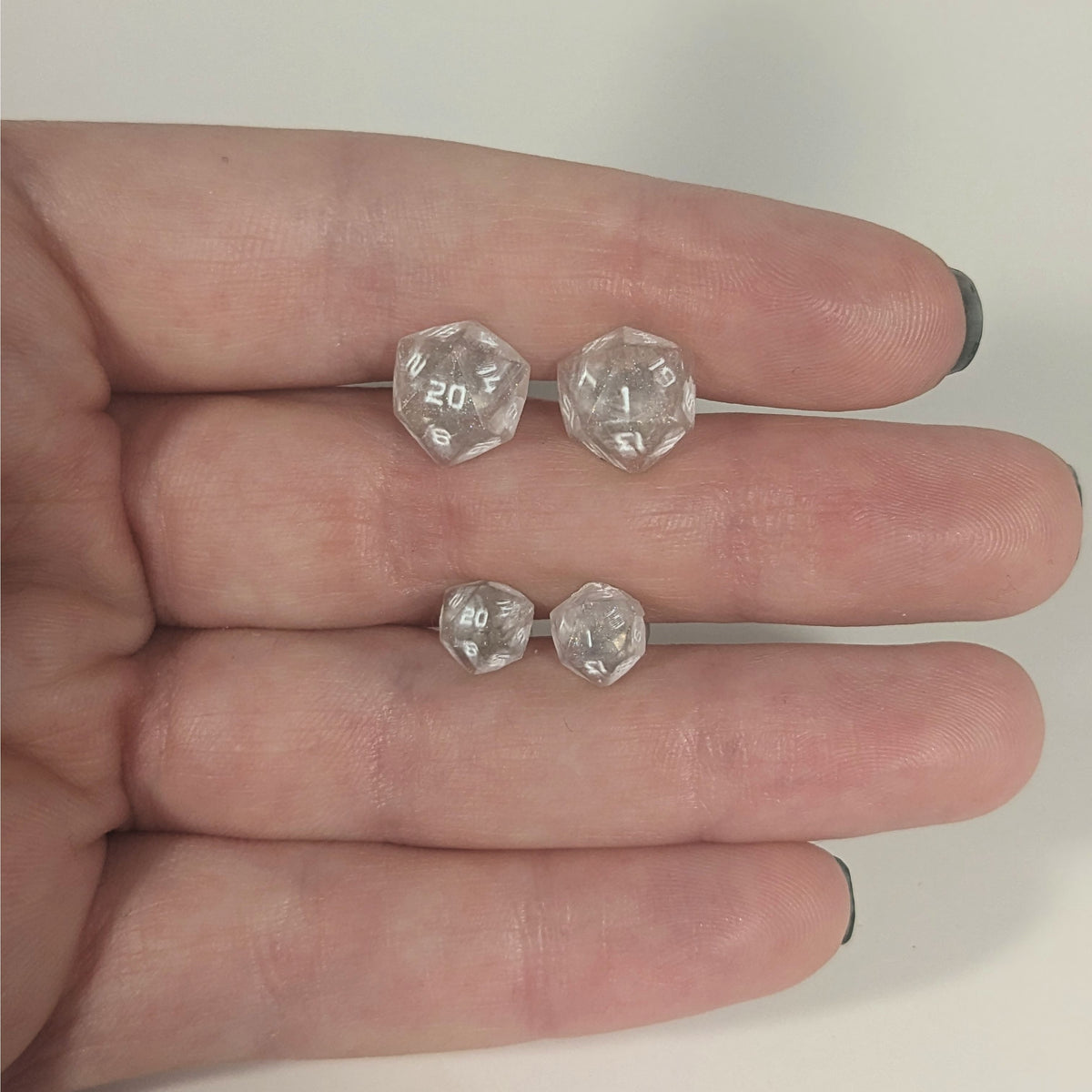 Two pairs of handmade d20 earrings with titanium hardware that are faded pink and translucent white with white numbers held between the fingers of a flat hand. The larger pair is between the pointer and middle finger, the smaller between the middle and ring finger. The earrings on the left of each pair centers the 20 face and the earrings on the right of each pair centers the 1 face.