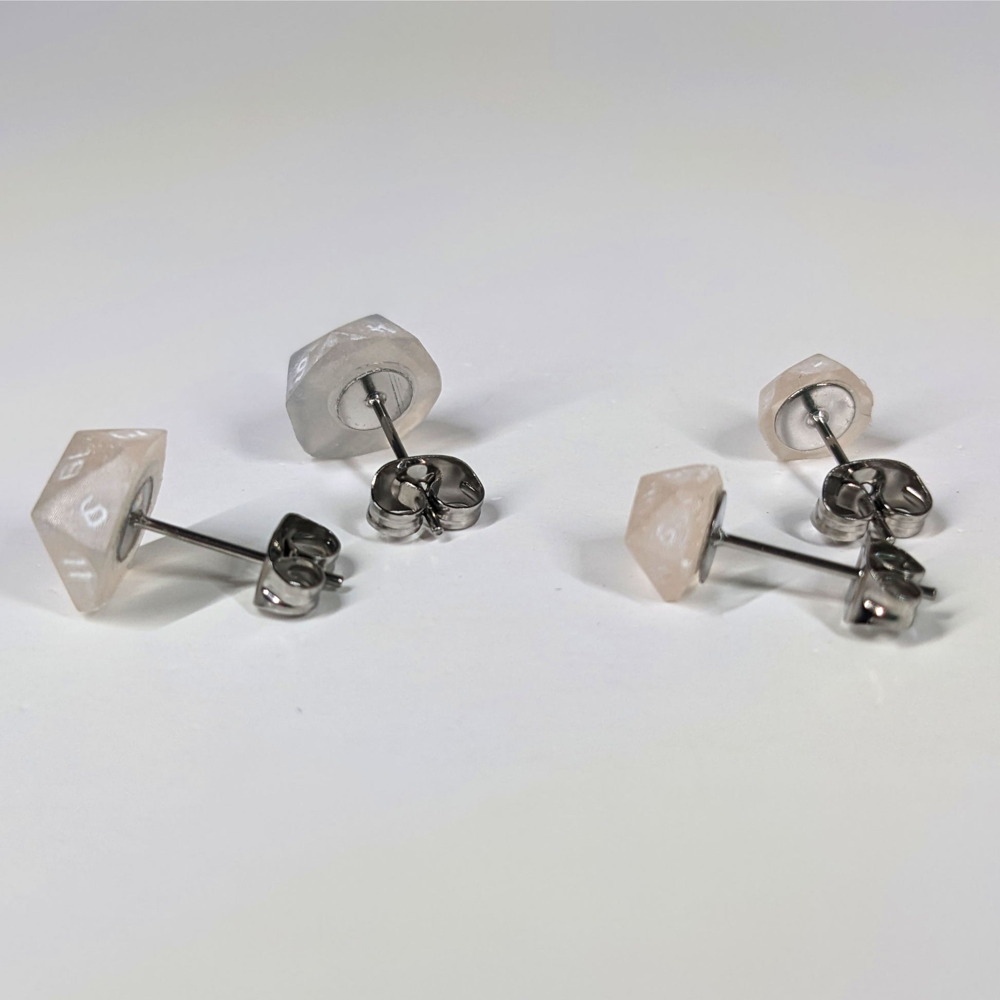 Two pairs of handmade d20 earrings that are light pink and white with white numbers turned to show the metal alloy hardware and butterfly backs.
