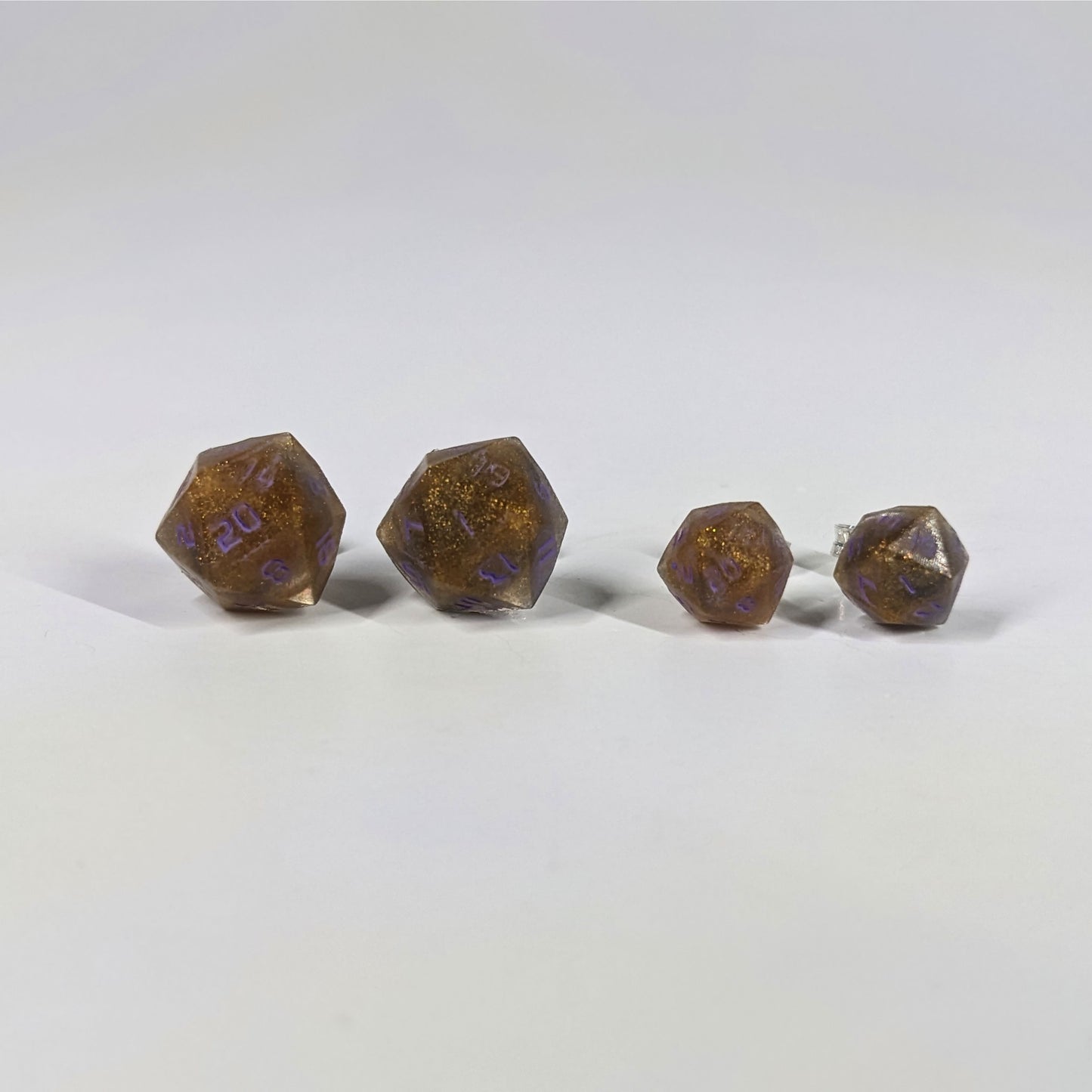 Two pairs of handmade d20 earrings with sterling silver hardware that are dark gold with purple numbers on a white background sit in a line with the larger pair on the left and the smaller pair on the right. The earrings on the left of each pair centers the 20 face and the earrings on the right of each pair centers the 1 face.